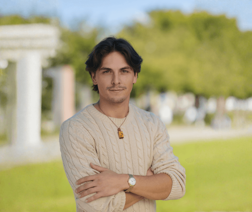 Young man wearing a beige cable-knit sweater standing with arms crossed in a park.