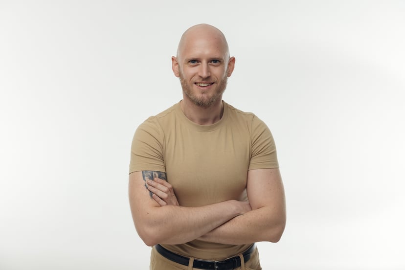 Smiling bald man with a beard and arm tattoo standing with arms crossed against a white background.