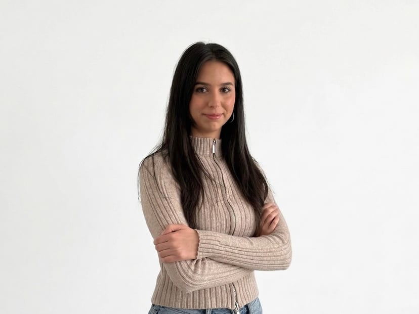 Young woman with long dark hair wearing a beige zip-up sweater standing with arms crossed against a white background.