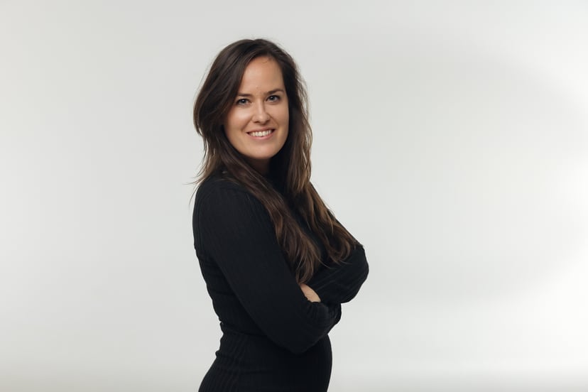Woman with long brown hair wearing a black long-sleeved shirt standing with arms crossed against a white background.