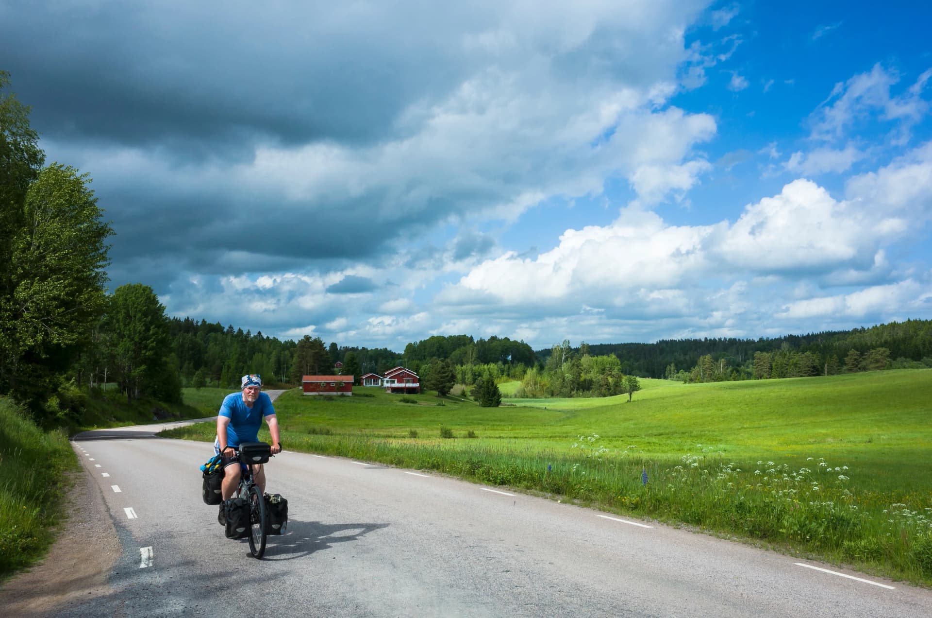 Cycling trip in Sweden, Man on touring bicycle solo travel Nordic countryside, Road among fields, gray clouds sunny summer day, Long distance cyclist travel around the world, Värmland