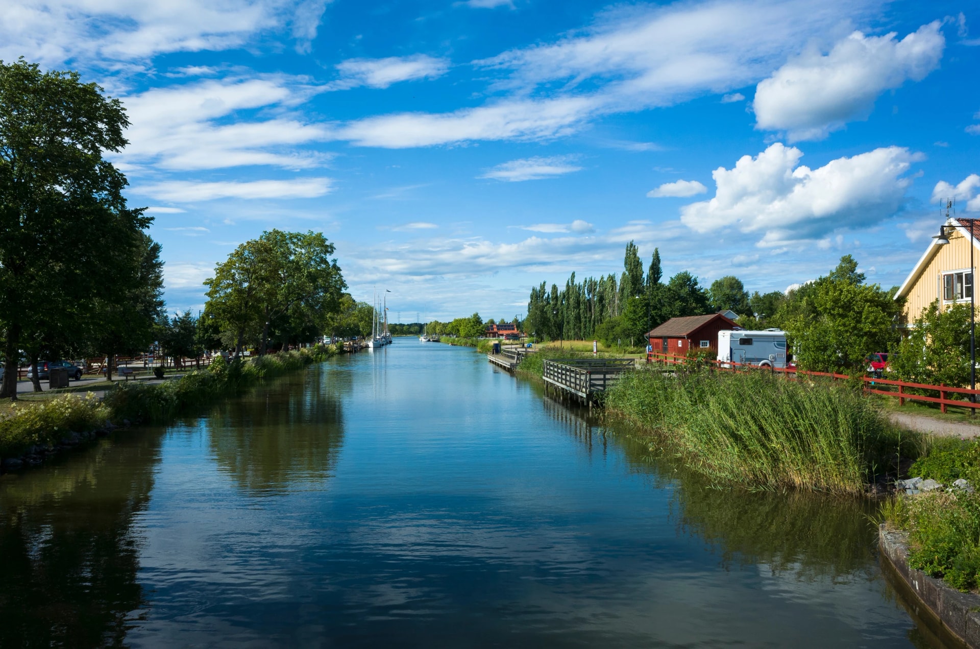 A scenic view of Göta Canal with calm waters reflecting a bright blue sky and white clouds. Lush greenery, traditional Swedish houses, and a peaceful summer atmosphere. Töreboda, Sweden