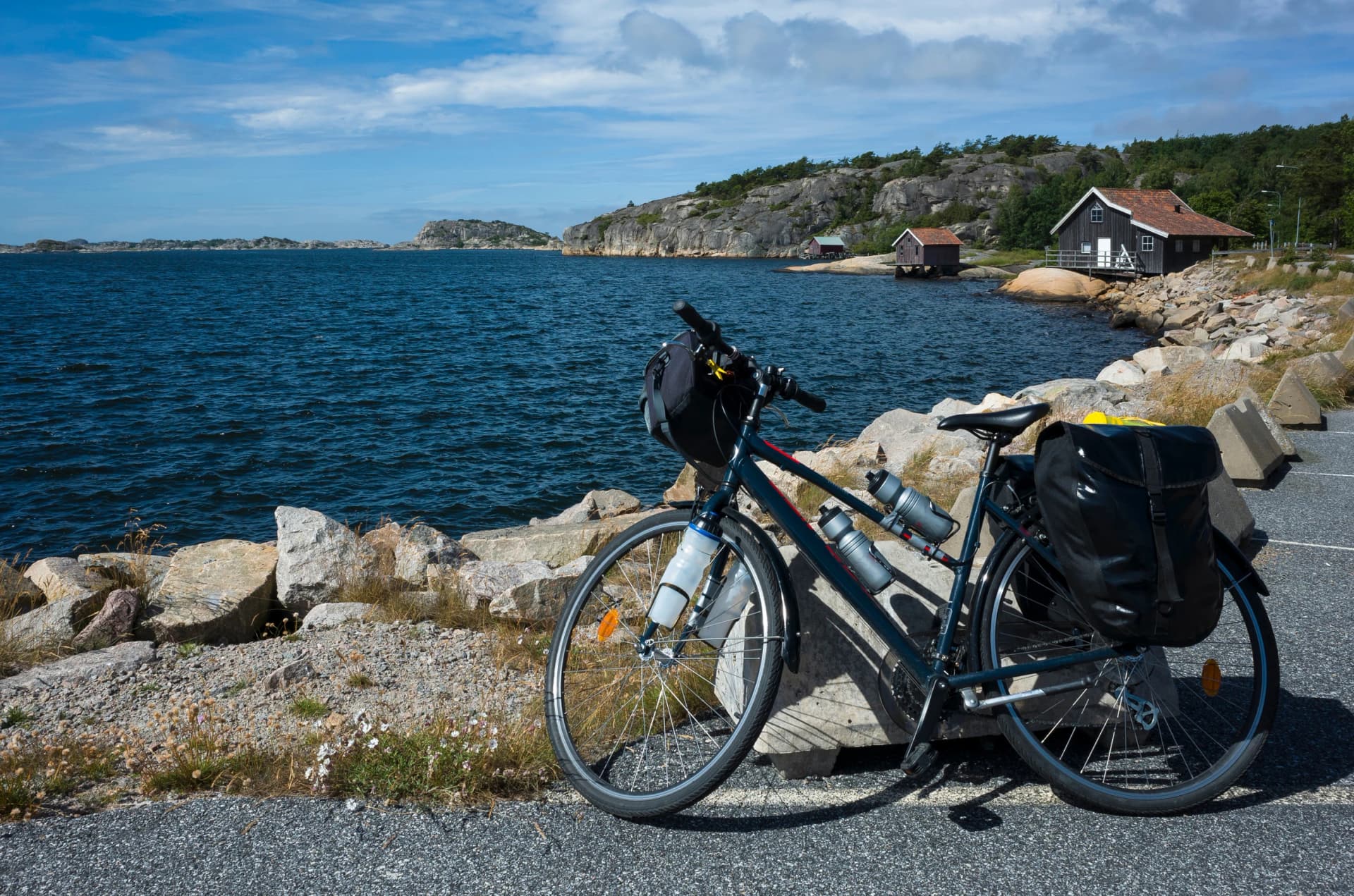 A touring bicycle with panniers leans against a rock at Kungsvik Harbor on Sweden’s west coast. A perfect summer stop on a cycling trip, with rocky shores, old boathouses, and blue water