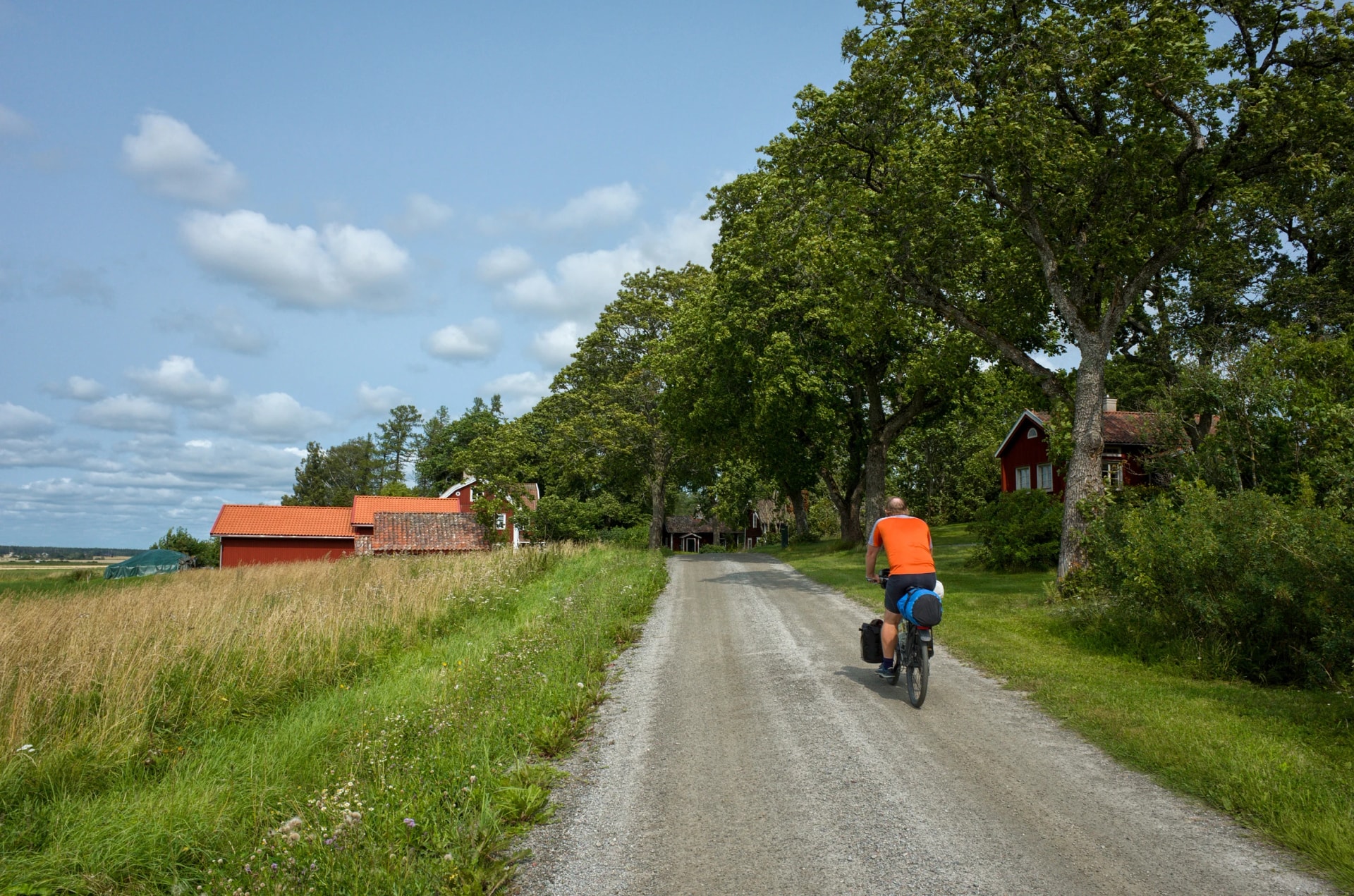 Cyclist on a gravel road in Västmanland, Sweden, passing red farm buildings and green fields under a partly cloudy summer sky