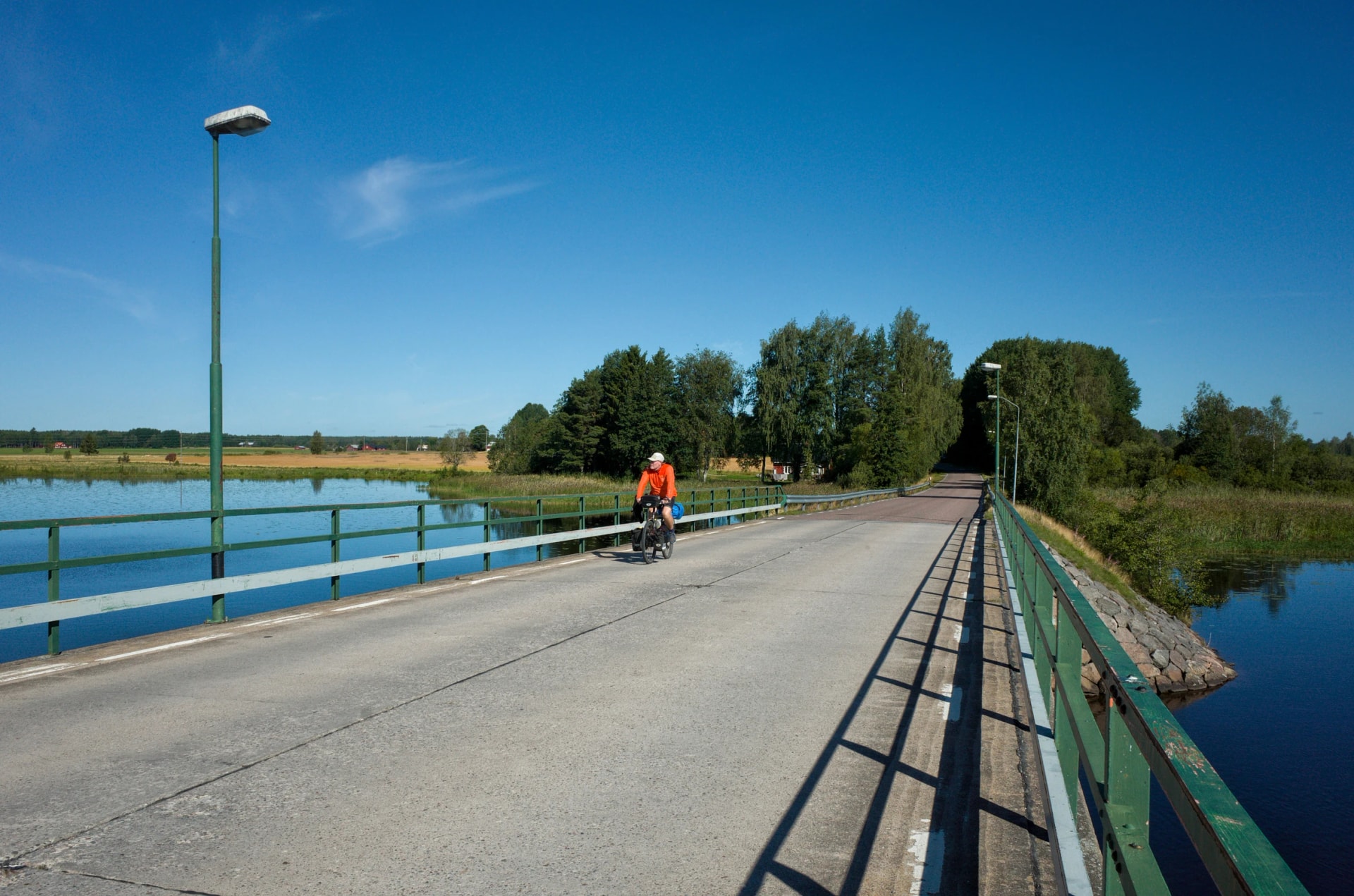 Cyclist crossing a bridge over the Dalälven river south of Hedesunda in Sweden, with calm water, green landscape, and clear blue sky